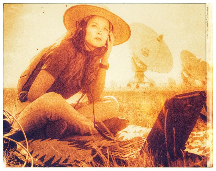 Ellie Aroway radio astronome assise un chapeau sur la tête et un casque audio sur les oreilles devant une rangée les énormes radio téléscopes du programme SETI.