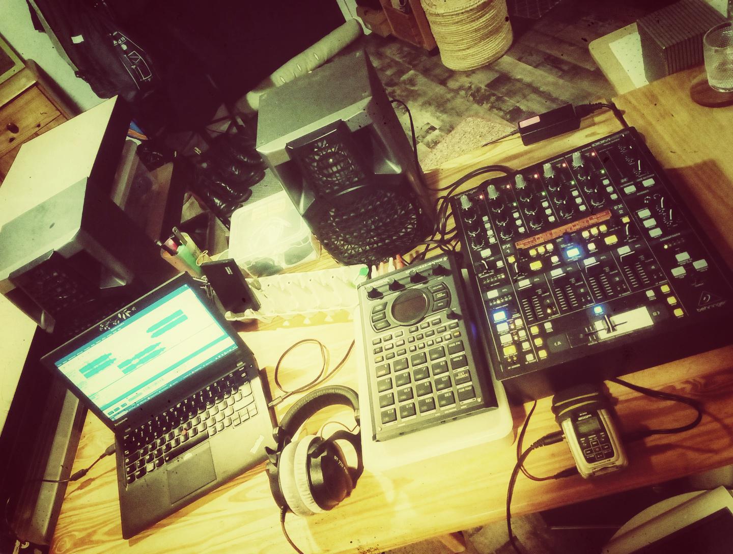 On voit une surface de travail sur laquelle est disposée un ordinateur portable dont l'écran affiche un logiciel d'édition audio. À côté est posé un casque audio, un sampleur Roland SP404 MKII et une table de mixage. Juste derrière il y a une paire d'enceinte.