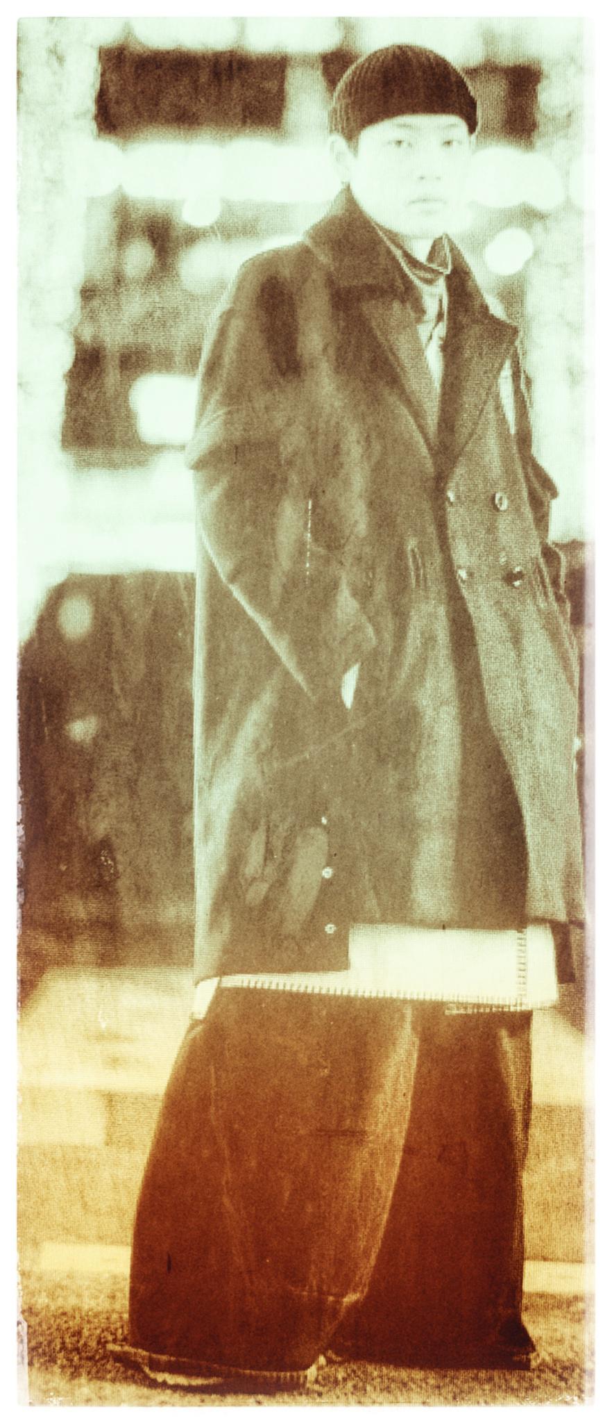 Une personne de type asiatique et queer, dans une rue, portant un long manteau et un pantalon large, regardant vers nous.