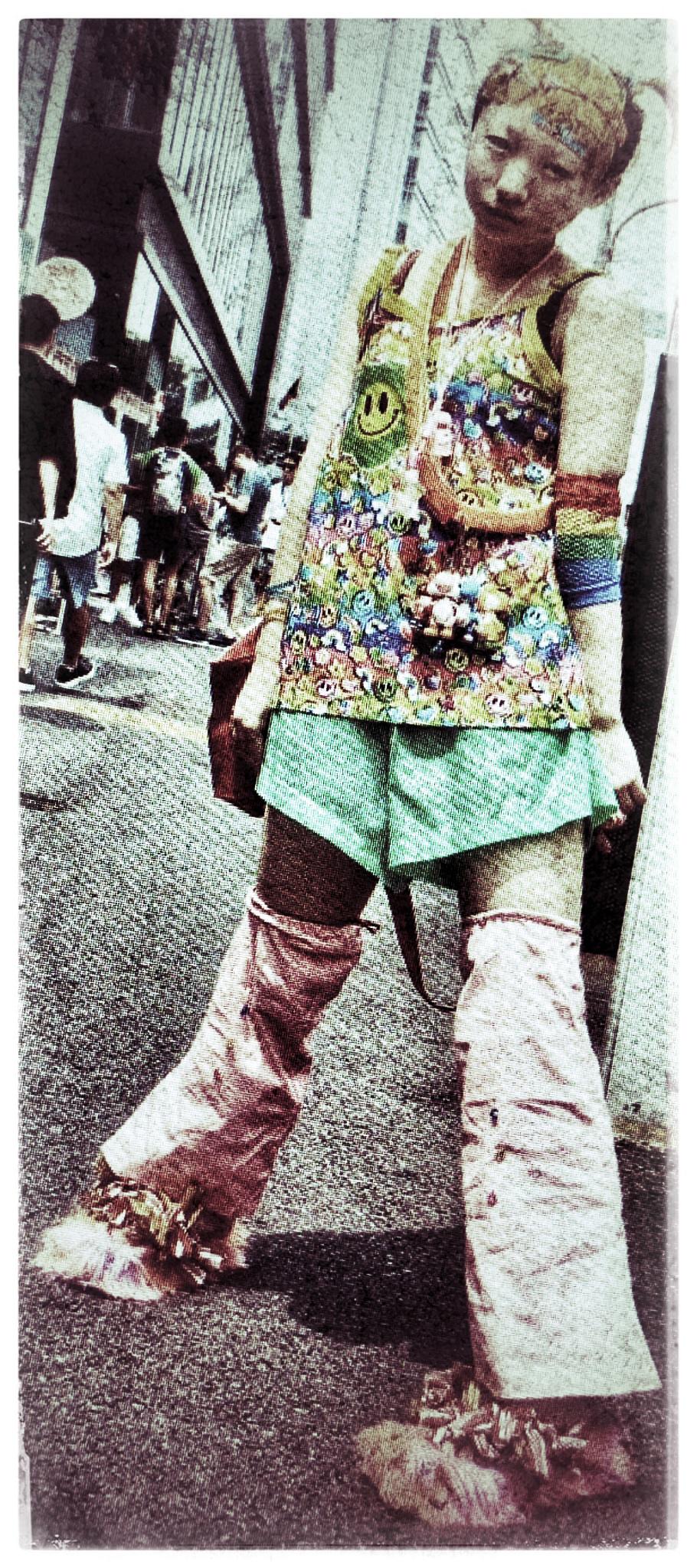 Une femme japonaise prise en photo dans une rue. Elle se tient debout les jamabes écartées. Elle est habillée de couches de vêtements multicolores .