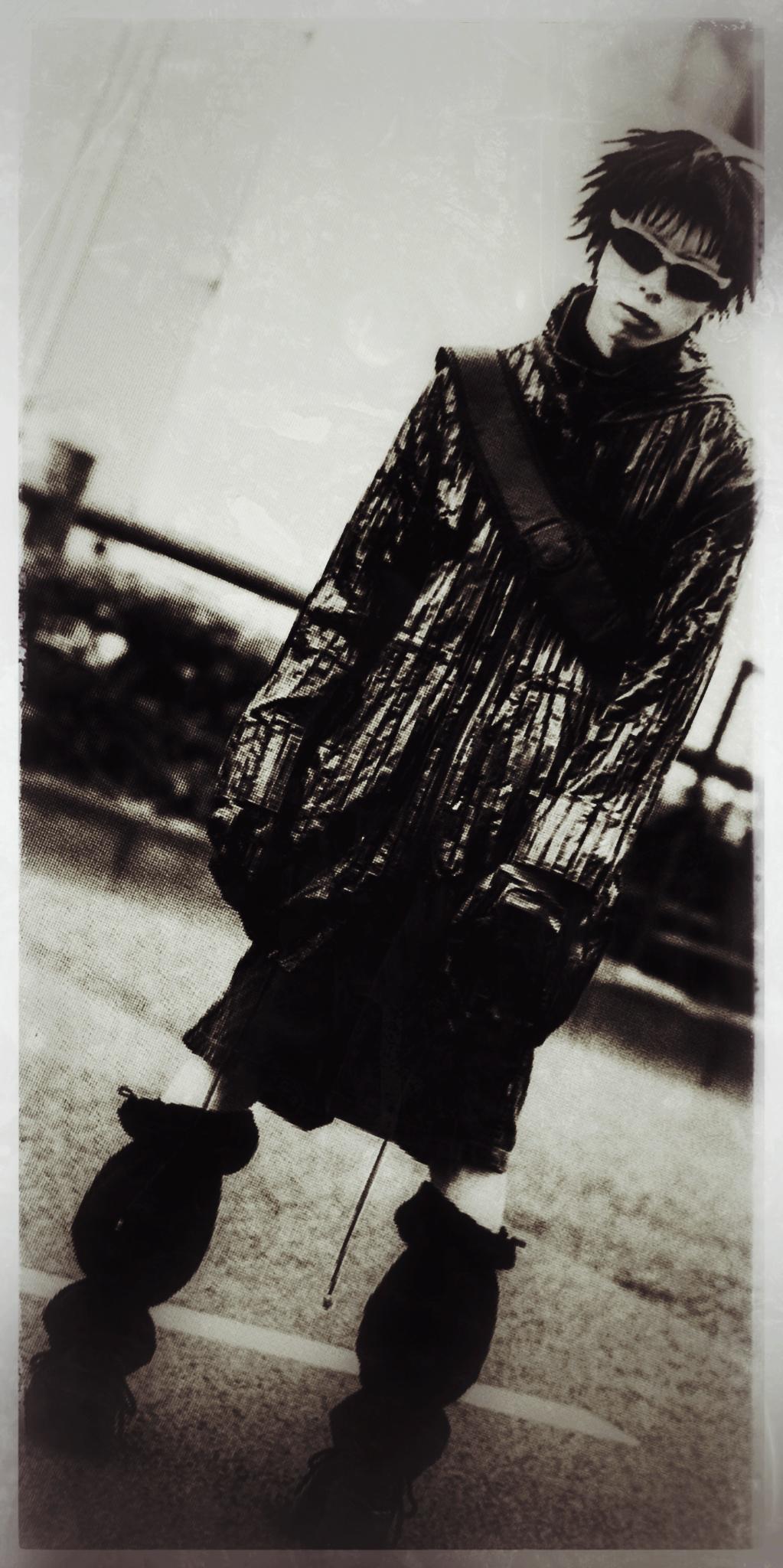 Un.e japonais.e queer habillé d'une jupe et d'une veste trois quart noires pris.e en photo dans une rue. Ielle regarde l'objectif.