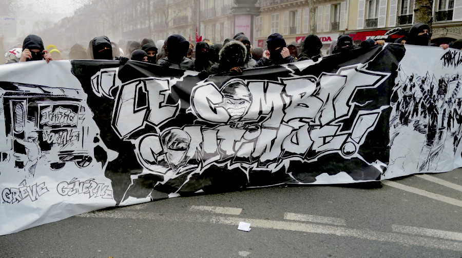 Scène de rue pendant un manifestation. Un cortège tend devant lui une large banderolle sur laquelle est écrit: "le combat continu"